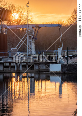 Sunset over industrial waterway with reflections and machinery under orange evening sky 114843777