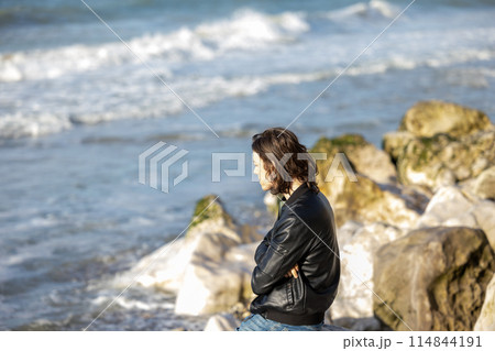 Picture of a contemplative woman sitting by the sea on a rocky beach, wearing a leather jacket Picture of a contemplative woman sitting by the sea on a rocky beach, wearing a leather jacket 114844191