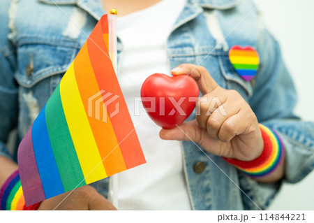 Asian lady wearing rainbow flag wristbands and hold red heart, symbol of LGBT pride month celebrate annual in June social of gay, lesbian, bisexual, transgender, human rights. Asian lady wearing rainbow flag wristbands and hold red heart, symbol of LGBT pride month celebrate annual in June social of gay, lesbian, bisexual, transgender, human rights. 114844221
