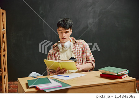 Young man, student sitting at desk and involved on book reading. Chalkboard background. Sitting in classroom and studying 114848068