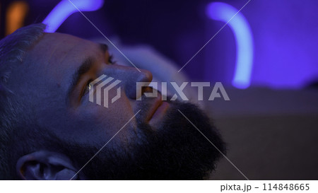 Close up side view of a bearded man looking up and thinking of something. Media. Young man with neon light on the background. 114848665
