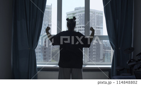 Rear view of a girl in casual clothes in modern bedroom in the morning. Media. Woman looking through the window, raising hands and stretching body muscles. Rear view of a girl in casual clothes in modern bedroom in the morning. Media. Woman looking through the window, raising hands and stretching body muscles. 114848802