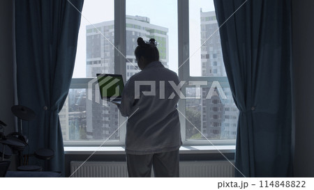 Rear view of woman in pajamas at home standing by the window and typing. Media. Young woman working on laptop with green screen. Rear view of woman in pajamas at home standing by the window and typing. Media. Young woman working on laptop with green screen. 114848822