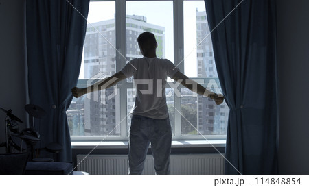 Early morning, silhouette of man exercising, stretching body by window at home. Media. Rear view of a young man standing against window. 114848854