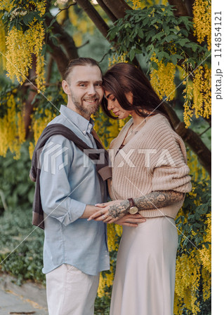 Amazing couple hugging under tree with yellow tree. Lovers couple hugging in a laburnum tree. Couple lovers hugging in the park. Girl with tattoos. A girl and a boy kiss. 114854121