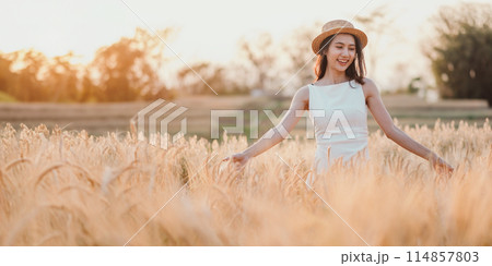 A woman is standing in a field of tall golden wheat. She is wearing a white dress and a straw hat. The scene is peaceful and serene, with the sun shining brightly overhead 114857803