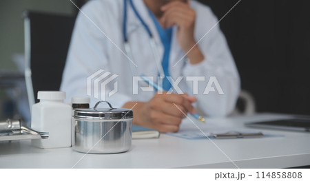 A professional and focused Asian female doctor in scrubs is working and reading medical research on her laptop in her office at a hospital. A professional and focused Asian female doctor in scrubs is working and reading medical research on her laptop in her office at a hospital. 114858808