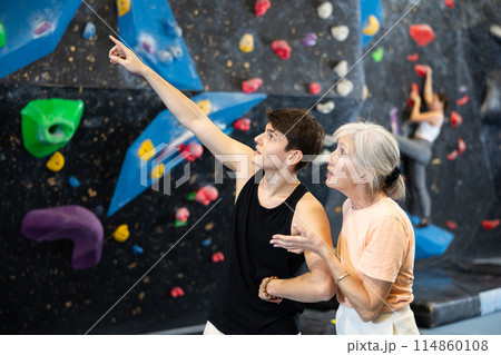 Young male trainer giving instructions to elderly woman about climbing artificial training rock wall in bouldering gym 114860108
