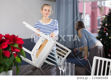Young woman dusts chair while an elderly mother vacuums room before Christmas 114860386