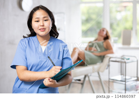 Smiling woman beautician in uniform standing in cosmetology clinic, filling out clipboard with medical records 114860397