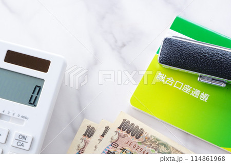 テーブルの上に置かれた銀行の預金通帳と印鑑、1万円札、電卓 テーブルの上に置かれた銀行の預金通帳と印鑑、1万円札、電卓 114861968