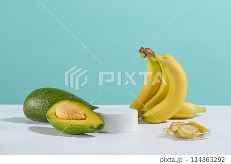 A small bunch of bananas and a few fresh avocados are scattered on white table around a circular empty platform in the middle, next to a glass dish with sliced banana against baby blue background A small bunch of bananas and a few fresh avocados are scattered on white table around a circular empty platform in the middle, next to a glass dish with sliced banana against baby blue background 114863292