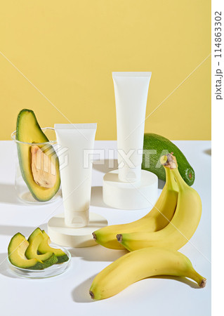 Design template with the theme of fruit ingredient for cosmetic product manufacturing, white table containing a cosmetic tube without label, banana and avocado decoration over yellow background 114863302