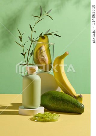 Sample cosmetic bottle in green color placed on a round platform with fresh avocado and yellow banana decorated in back, a dish of chopped avocado placed in front. Green background photo, front view 114863319