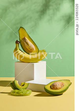 The yellow flatform containing a white platform with a banana bunch and a fresh avocado above, next to a blank podium, decorated with some fresh avocado in front over green background 114863320