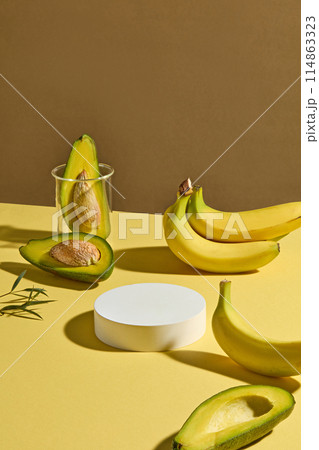 The creative design with a yellow countertop containing a blank white podium in center and some yellow banana and fresh avocado flat lay above striking on brown background, space for adding elements 114863323
