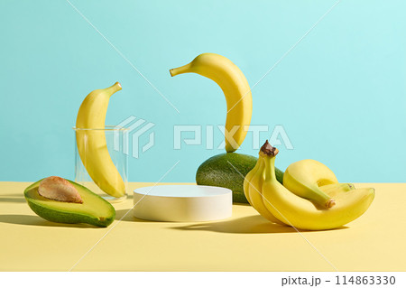 Baby blue background against several avocado and banana displayed on the yellow flat surrounded a white podium in center for showing product. Photo for promotion with displaying space 114863330