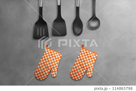 Orange heat resistant cooking gloves with kitchen utensils hanging on cement wall. Orange heat resistant cooking gloves with kitchen utensils hanging on cement wall. 114863798