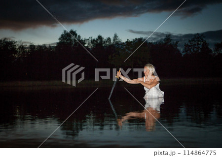 A woman in a white dress stands in a lake with a candle at night, creating a serene atmosphere 114864775