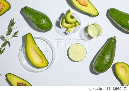 White background with view from above features avocado cut in halves and slices. A beaker and a petri dish containing avocado cream. Copy space 114866047