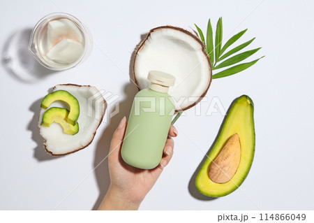 Flat lay of hand model holding a pastel green bottle without label. Fresh coconuts and avocados decorated over white background. Skincare concept with empty label 114866049