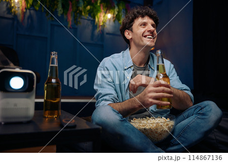 Smiling man with popcorn and beer watching movie on projector Smiling man with popcorn and beer watching movie on projector 114867136
