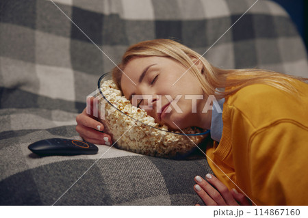 Sleepy woman is resting holding her head in plate with popcorn Sleepy woman is resting holding her head in plate with popcorn 114867160