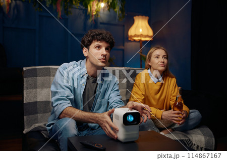 Man and a woman are sitting on a couch watching a movie on a projector Man and a woman are sitting on a couch watching a movie on a projector 114867167