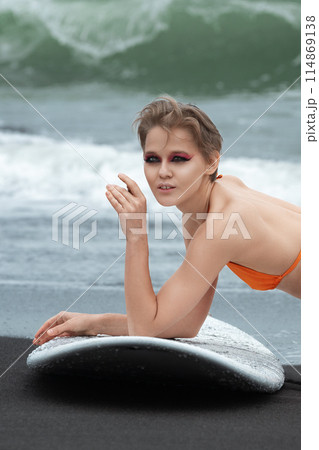 Sexy surfer lying on black sandy beach with surfboard, showcasing fit and athletic body. Caucasian sportswoman is relaxing after sports training session. Sports activities, joy they bring to our lives 114869138