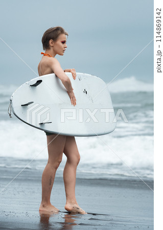 Gorgeous surfer woman stands on black, sandy beach, carrying surfboard against backdrop of rolling sea waves. Woman tourist, full of joy of summer vacation, looks on at magnificent Pacific Ocean waves Gorgeous surfer woman stands on black, sandy beach, carrying surfboard against backdrop of rolling sea waves. Woman tourist, full of joy of summer vacation, looks on at magnificent Pacific Ocean waves 114869142