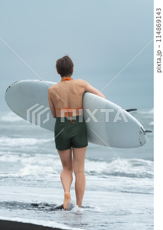 Rear view of female surfer on summer beach carrying her board and looking like an unstoppable force as she strides along sand. It's great picture of woman enjoying sports training during beach holiday Rear view of female surfer on summer beach carrying her board and looking like an unstoppable force as she strides along sand. It's great picture of woman enjoying sports training during beach holiday 114869143