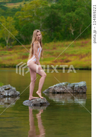 Sexy young woman poses for photo in nature, stands on rocky island in lake surrounded by colorful autumn forest. Beautiful model wearing one piece swimsuit. Photo captures her sensuality and beauty Sexy young woman poses for photo in nature, stands on rocky island in lake surrounded by colorful autumn forest. Beautiful model wearing one piece swimsuit. Photo captures her sensuality and beauty 114869221