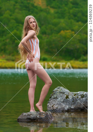 Young woman with long blonde hair is standing on rocky island in lake surrounded by colorful autumn forest. Sexy lady wearing one piece swimsuit and appears to be tourist enjoying beautiful scenery 114869239