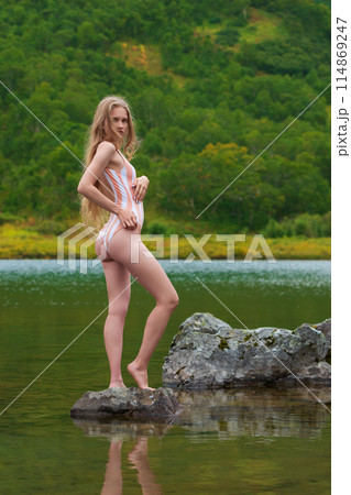 Woman standing on rocky island in middle of lake surrounded by most beautiful autumn forest. Blonde woman wearing one piece swimsuit who's having time of her life enjoying breathtaking nature around 114869247