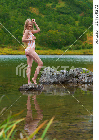 Serene woman in one piece swimsuit with long blonde hair is standing on rocky island in lake, surrounded by colorful autumn forest. Beautiful young Caucasian woman is tourist, relaxing in nature Serene woman in one piece swimsuit with long blonde hair is standing on rocky island in lake, surrounded by colorful autumn forest. Beautiful young Caucasian woman is tourist, relaxing in nature 114869272