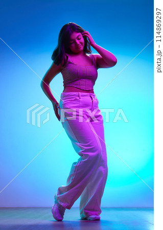 Young Indian woman in stylish attire dancing of joy and happiness in pink neon light against gradient blue background. 114869297