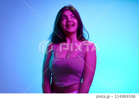 Young Indian girl, 20s years old, looking away with playful expression in pink neon light against gradient blue background. 114869386