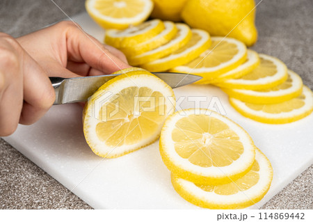 Woman cutting lemon into slices with a sharp utility knife on a stone kitchen board. Woman cutting lemon into slices with a sharp utility knife on a stone kitchen board. 114869442