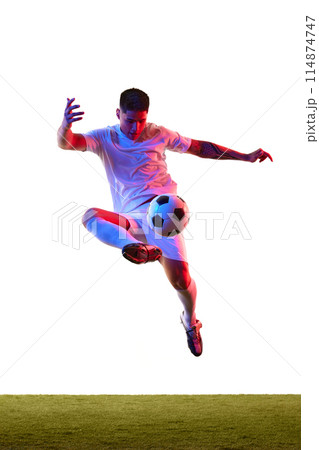 Dynamic shot of young guy dressed sportswear kicking ball in motion in neon light against white studio background. Dynamic shot of young guy dressed sportswear kicking ball in motion in neon light against white studio background. 114874747