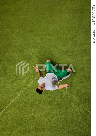 Moment of triumph or celebration. Aerial view of soccer player seated on green grass and gesturing radiating excitement. 114874788