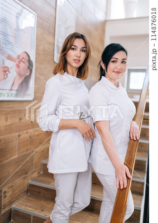 Vertical portrait of two young female practitioner doctors in medical uniform smiling looking at camera standing posing on staircase in medic clinic. Portrait of medical staff posing on hospital. Vertical portrait of two young female practitioner doctors in medical uniform smiling looking at camera standing posing on staircase in medic clinic. Portrait of medical staff posing on hospital. 114876018