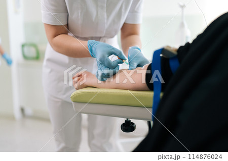 Nurse gives intravenous infusion to female patient in clinic. Young medical worker in scrubs uniform inserts IV line needle in vein of woman sitting on armchair at hospital. Medicine, vitamin therapy. 114876024