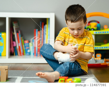 A little toddler boy holds his leg in a cast with his hands. Fracture of a foot and finger in children. Human healthcare and medicine concept. Bandage A little toddler boy holds his leg in a cast with his hands. Fracture of a foot and finger in children. Human healthcare and medicine concept. Bandage 114877391