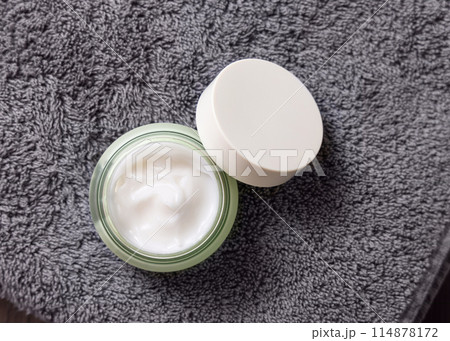 Opened cream jar with blank white lid on dark grey towel top view, cosmetic mockup 114878172