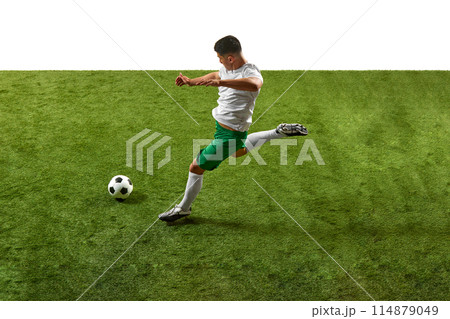 Action shot of young athletic guy, footballer in green and white gear, executing powerful kick on grassy pitch isolated white backdrop. 114879049