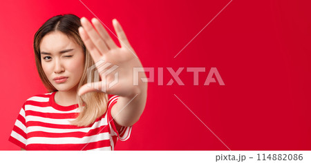Close-up serious-looking determined asian blond woman pull hand forward show stop taboo sign, concentrating gaze on camera look with one eye, demanding stop, tell enough, say no, red background Close-up serious-looking determined asian blond woman pull hand forward show stop taboo sign, concentrating gaze on camera look with one eye, demanding stop, tell enough, say no, red background 114882086