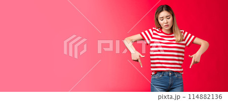 Disappointed gloomy blond asian girl sighing upset looking pointing fingers down, express regret sorrow, stand red background wear striped t-shirt, whining wish not come true 114882136