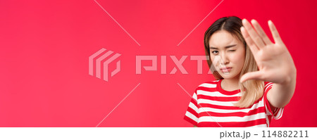 Close-up serious-looking determined asian blond woman pull hand forward show stop taboo sign, concentrating gaze on camera look with one eye, demanding stop, tell enough, say no, red background Close-up serious-looking determined asian blond woman pull hand forward show stop taboo sign, concentrating gaze on camera look with one eye, demanding stop, tell enough, say no, red background 114882211