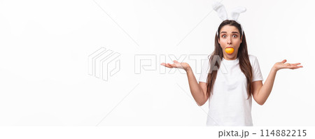 Portrait of confused and indeisive young funny woman with rabbit ears, holding colored Easter egg in mouth, staring clueless camera with popped eyes, shrugging and spread hands sideways Portrait of confused and indeisive young funny woman with rabbit ears, holding colored Easter egg in mouth, staring clueless camera with popped eyes, shrugging and spread hands sideways 114882215