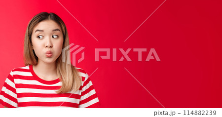 Close-up silly tender young modern asian girl blond haircut hold breath, folding lips and pouting look away as if hiding secretn, stand red background innocent clueless seal lips with promise 114882239